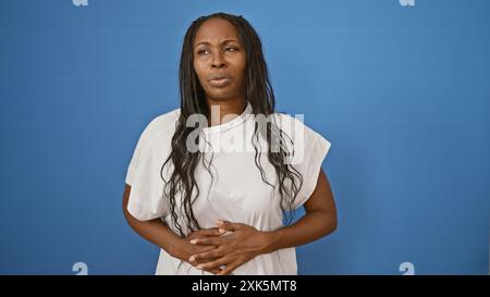 Une jeune femme afro-américaine adulte avec des cheveux bouclés se tient sur un fond bleu, tenant son estomac dans l'inconfort. Banque D'Images