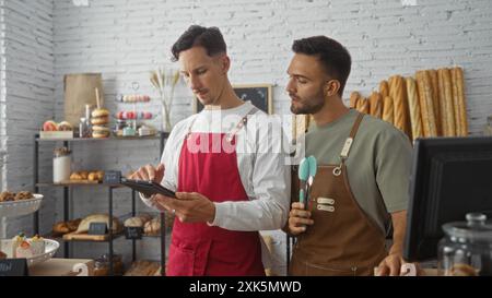 Deux hommes professionnels adultes, ouvriers dans une boulangerie, discutent des tâches ensemble tout en utilisant une tablette dans un cadre intérieur bien éclairé rempli de divers bre Banque D'Images