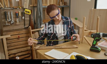 Une femme concentrée mesure le bois avec un ruban adhésif dans un atelier, entourée d'outils et d'un établi. Banque D'Images