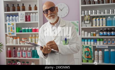 Homme d'âge moyen aux cheveux gris et à la barbe, portant des lunettes et une blouse de laboratoire, prenant des notes dans une pharmacie bien approvisionnée. Banque D'Images