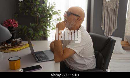 Homme assis dans la chambre à coucher, travaillant de la maison sur ordinateur portable, avec une expression réfléchie, dans un cadre intérieur confortable et élégant avec des plantes d'intérieur et un décor, seni Banque D'Images