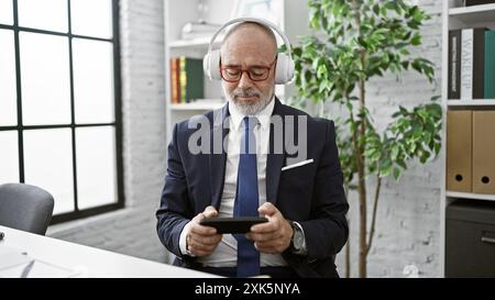 Un homme mûr en costume aime la musique avec des écouteurs dans un bureau moderne, tenant une tablette. Banque D'Images