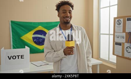 Beau jeune homme afro-américain avec une barbe tenant un micro, debout dans une salle de collège électoral avec un drapeau brésilien en arrière-plan. Banque D'Images