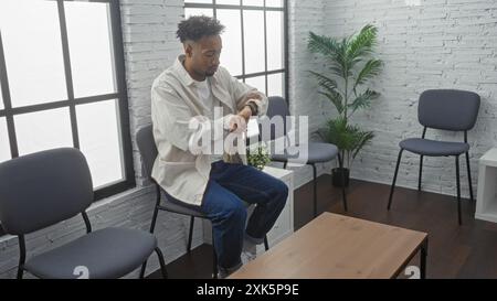 Jeune homme afro-américain avec une barbe vérifiant sa montre tout en étant assis dans une salle d'attente intérieure au décor moderne, avec des murs de briques blanches et Banque D'Images