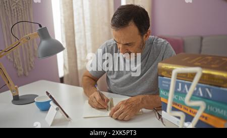 Homme hispanique d'âge moyen écrivant dans un cahier à la maison avec des livres sur un bureau, se concentrant intensément dans un salon très éclairé. Banque D'Images