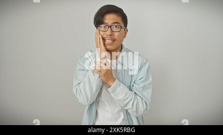Un jeune homme asiatique pensif avec des lunettes posant devant un fond blanc, habillé décontracté dans une veste en denim léger. Banque D'Images