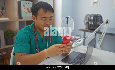 Un jeune vétérinaire chinois dans une clinique portant des gommages verts et un stéthoscope regarde son smartphone près d'un ordinateur portable. Banque D'Images