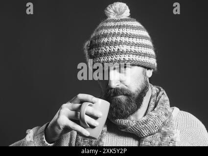 Sérieux homme élégant en pull et chapeau enveloppé dans une écharpe avec tasse de café ou de thé. Automne ou hiver. Portrait d'homme barbu à la mode Banque D'Images