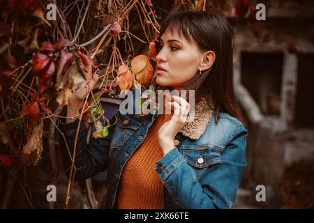 Une jeune femme aux longs cheveux foncés, vêtue d'une veste en Jean et d'un col roulé orange, pose devant un vieux bâtiment, entouré de feuilles d'automne. Banque D'Images