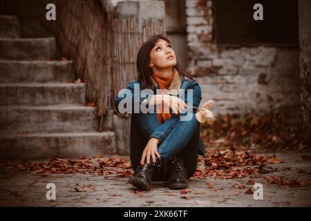 Une jeune femme est assise sur des marches de pierre, entourée de feuilles tombées, regardant le ciel. Banque D'Images