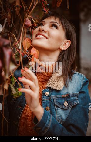Une jeune femme aux longs cheveux bruns sourit à une branche de feuilles d'automne tout en portant une veste en denim. Banque D'Images