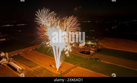 Des feux d'artifice brillants éclatent dans le ciel nocturne, illuminant Un parc d'expositions rural ci-dessous avec de la lumière dorée et blanche, entouré de champs sombres et D'Une atmosphère bondée et festive. Banque D'Images