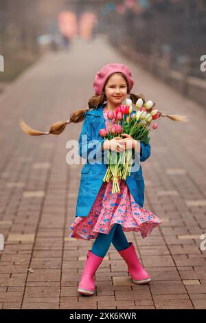 Belle fille enfant avec bouquet de fleurs de tulipes roses de printemps à la rue de la ville. Portrait de jolie enfant dans le parc en plein air. Banque D'Images