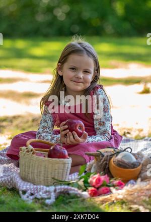 Portrait d'une jolie fille mignonne sur un pique-nique. Un enfant tient une pomme rouge dans ses mains et sourit ; un panier de fruits se trouve à proximité. Banque D'Images