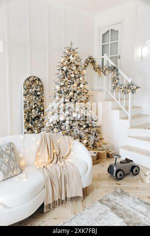 Un élégant canapé en cuir blanc sur la toile de fond d'un grand sapin de Noël à l'intérieur d'un salon blanc avec un escalier en bois menant à Banque D'Images