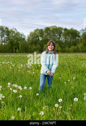 Une petite fille heureuse rit joyeusement et se tient sur un champ vert avec des pissenlits moelleux, recueillant joyeusement les peluches des fleurs. Enfants et nat Banque D'Images