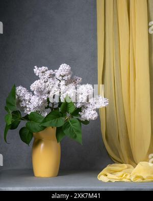 Un grand bouquet de lilas blancs dans un vase jaune sur un fond gris, avec un rideau jaune suspendu à proximité, espace pour le texte. Nature morte avec des lilas Banque D'Images