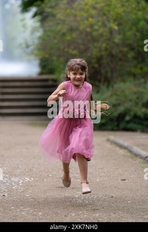 Une petite fille mignonne dans une belle robe rose avec de longs cheveux court rapidement. Jeux de plein air. Arrière-plan flou. Banque D'Images