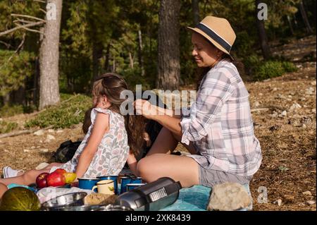 Mère tressant les cheveux de sa jeune fille tout en profitant d'un pique-nique familial relaxant dans un cadre forestier tranquille. Les pommes et un thermos ajoutent au confortable a Banque D'Images