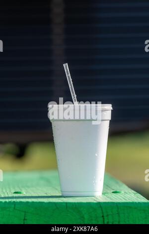 Un grand gobelet blanc à emporter non recyclable avec un couvercle en plastique et de la paille sur une table de patio vert vif. Le gobelet en polystyrène ou en styromousse est isolé Banque D'Images