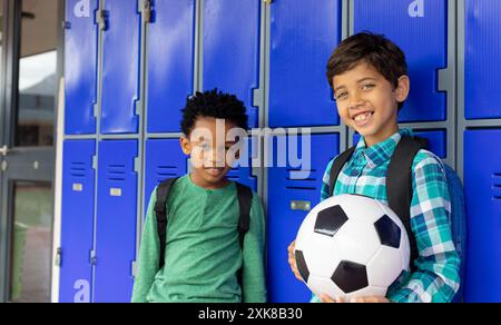 Garçon biracial et garçon afro-américain se tiennent souriant près des casiers de l'école, l'un tenant un ballon de football Banque D'Images