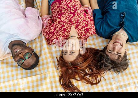 Vue aérienne de divers groupes de jeunes amis allongés sur une couverture de pique-nique jaune à carreaux. Jeunesse multiculturelle profitant de la journée d'été à l'extérieur, souriant an Banque D'Images