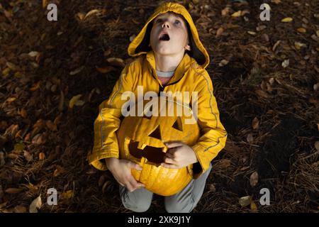 Enfant effrayé dans une veste orange est assis sur le sol parsemé de feuilles mortes, tenant une grande citrouille d'Halloween Banque D'Images