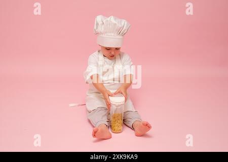 Un chef enfant montre fièrement un pot de pâtes crues sur un fond de studio rose. Enfant âgé de 3 ans (garçon de 3 ans) Banque D'Images