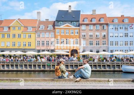 Deux personnes s'assoient sur une passerelle pavée le long d'un canal à Copenhague, profitant du soleil et d'une vue sur les bâtiments colorés. Copenhague Danemark Banque D'Images