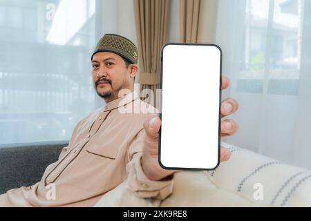 Heureux arabe montrant un téléphone portable avec un écran vide. Heureux homme musulman asiatique montrant un écran blanc blanc de téléphone. Jeune homme islamique assis sur le canapé Banque D'Images