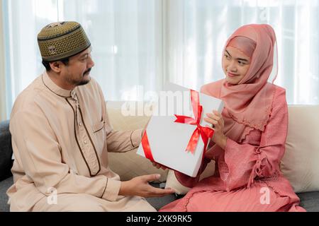 Mari musulman donnant une boîte-cadeau emballée à la femme surprise célébrant les vacances de Noël ou la Saint-Valentin ensemble assis sur le canapé à la maison. Vacances ce Banque D'Images