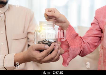 Couple musulman avec pot de pièces de monnaie. Main de femme mettant de l'argent dans le bocal en verre étiqueté tenu par un homme, économiser et investir de l'argent pour se préparer à l'avenir Banque D'Images