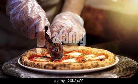 Un chef découpe habilement une pizza chaude sur un plateau en métal dans une cuisine professionnelle. Le couteau tranche à travers la création recouverte de mozzarella, la rendant prête t Banque D'Images