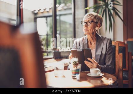Femme mature est assise dans le café et relaxant. Elle boit du café et utilise une tablette numérique. Banque D'Images