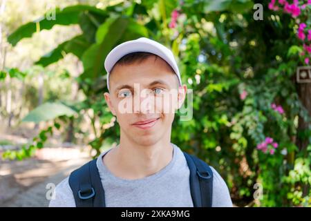 Jeune homme avec une casquette blanche et un sac à dos souriant à la caméra dans un parc vert luxuriant, portant un t-shirt gris. Concept d'aventure en plein air Banque D'Images