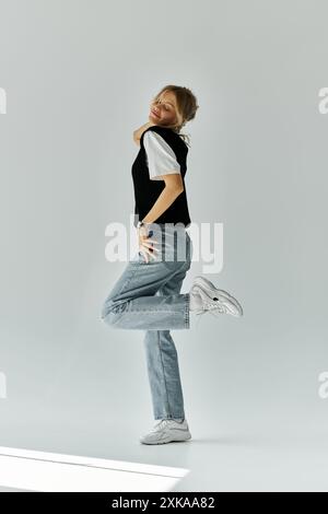 Une jeune femme aux cheveux blonds frappe une pose dans une chemise noire et un Jean sur un fond gris. Banque D'Images