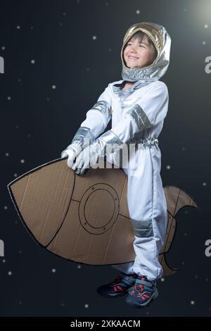 Un enfant souriant dans une combinaison spatiale chevauchant une fusée en carton voyageant dans l'espace. Banque D'Images