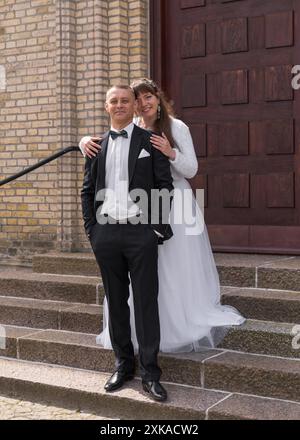 La mariée et le marié sont debout sur les marches contre la toile de fond des portes brunes. Le marié garde ses mains dans ses poches. La mariée se tient derrière t Banque D'Images