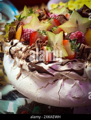 Gros plan d'un dessert pavlova garni de fruits frais, dont des fraises, des framboises, des fruits étoilés et des copeaux de chocolat. Banque D'Images