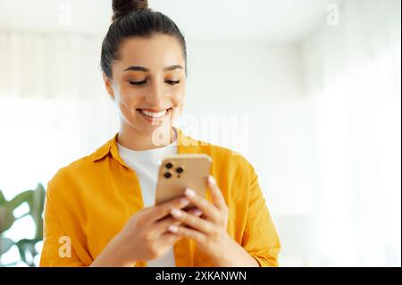 Utilisation de gadgets. Photo d'une jeune femme positive jolie arabe ou indienne en chemise orange tenant un smartphone dans les mains, tapant un message dans les médias sociaux, naviguant sur Internet, vérifiant les e-mails Banque D'Images
