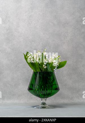 Fleurs blanches, lys de la vallée dans un vase vert transparent, un verre, à côté se trouve une écharpe verte et trois boules vertes. Fond gris. Beaucoup Banque D'Images