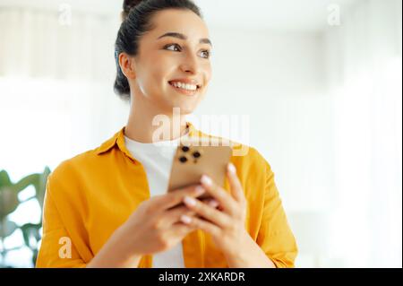 Photo en gros plan d'une belle jeune femme arabe ou indienne positive en chemise orange tenant un smartphone dans les mains, tapant un message dans les médias sociaux, naviguant sur Internet, commandant des produits en ligne dans des applications Banque D'Images