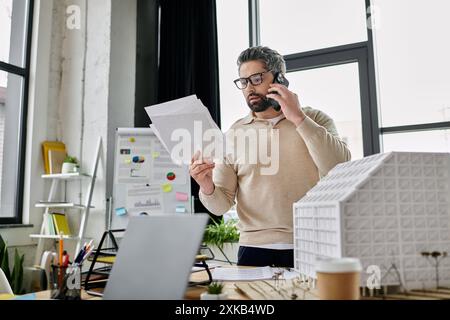 Un bel homme d'affaires avec une barbe travaille dans un bureau moderne. Il est au téléphone et examine des documents tout en se tenant debout à côté d'un bâtiment modèle. Banque D'Images