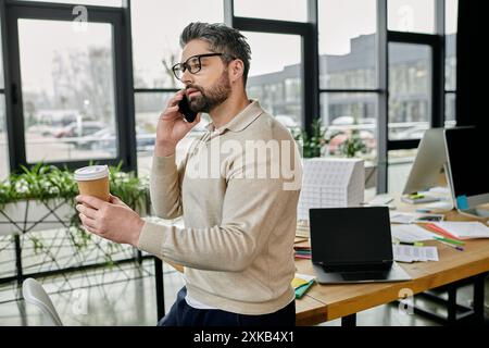 Un bel homme d'affaires avec une barbe, portant des lunettes, se tient dans un bureau moderne tout en tenant un café et en parlant au téléphone. Banque D'Images