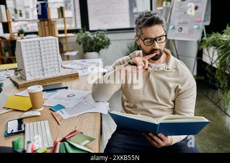 Un bel homme d'affaires avec une barbe est assis dans un bureau moderne, tenant soigneusement un stylo à son menton tout en lisant un livre. Banque D'Images