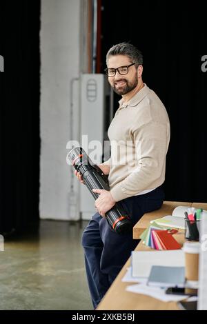 Un bel homme d'affaires avec une barbe est assis à un bureau moderne, tenant un tube et souriant avec confiance. Banque D'Images