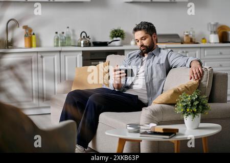 Un homme barbu dans une chemise décontractée et un pantalon se détend sur un canapé dans un cadre de maison, à l'aide de son smartphone. Banque D'Images