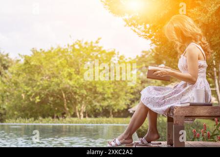 Jeune femme dans une robe blanche vintage est assise sur une terrasse au bord de la piscine lisant la Bible seule pour apprendre et comprendre les enseignements de Dieu de la Bible qu'elle est Banque D'Images