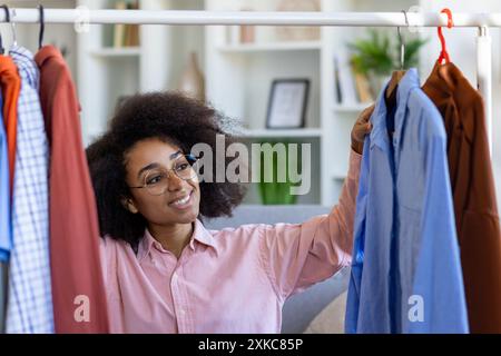 Femme souriante sélectionnant des chemises de la garde-robe et décidant quoi porter. Profitez de l'organisation et de la création de tenues élégantes à la maison. Intérieur moderne de la maison en arrière-plan. Banque D'Images