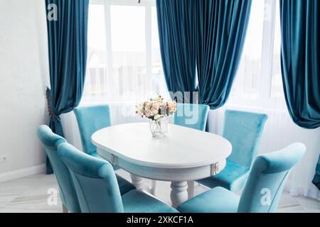 Lumineuse et élégante salle à manger avec rideaux et chaises en velours bleu, une table blanche et une pièce maîtresse florale. Banque D'Images